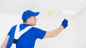 Malarz w niebieskim uniformie maluje sufit białym wałkiem; widoczne ślady po taśmie malarskiej.