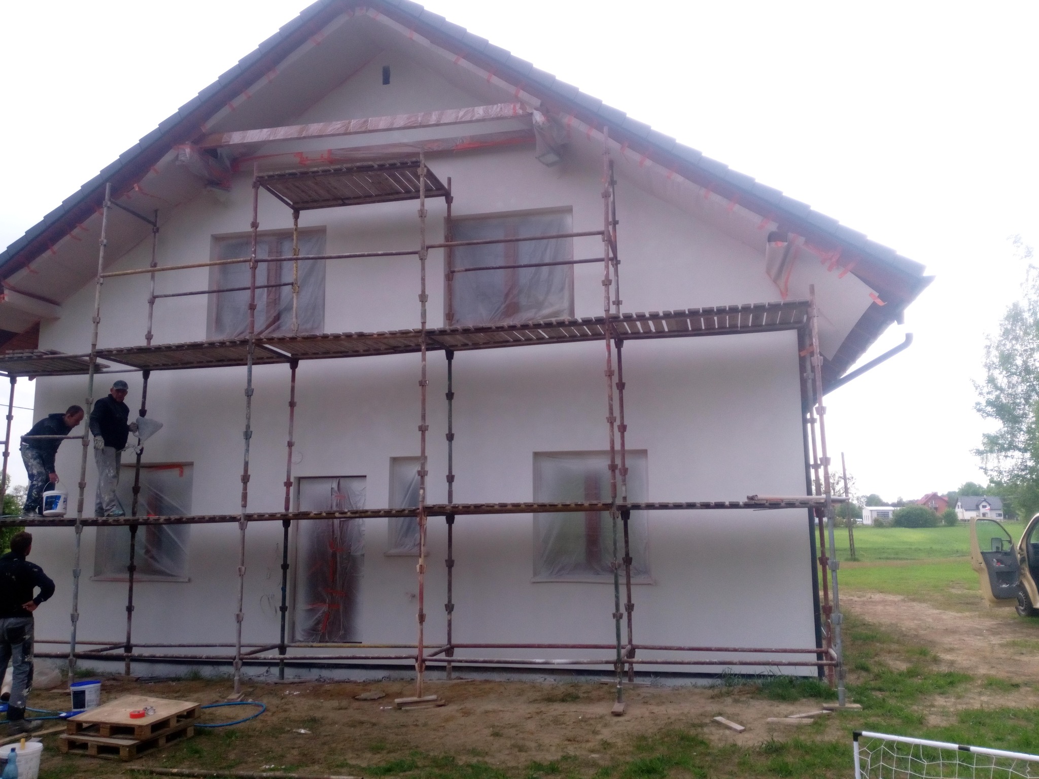 Prace tynkarskie na elewacji domu jednorodzinnego, dwóch pracowników na rusztowaniu tynkuje elewację, okna zabezpieczone folią, widoczny fragment działki i samochód z otwartymi drzwiami.