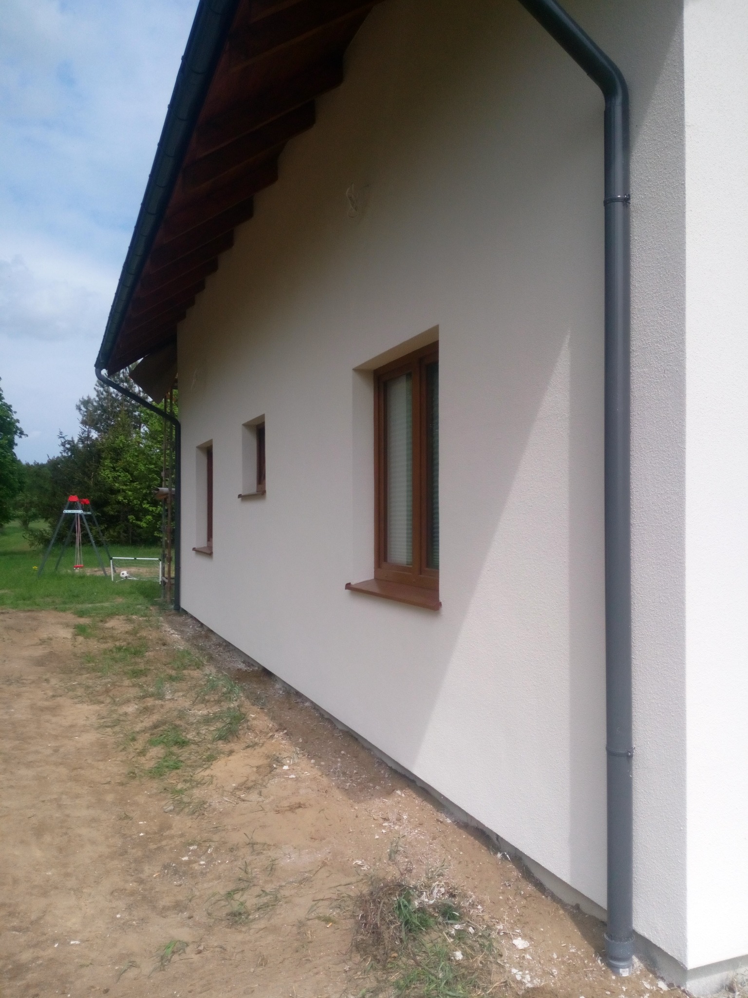 Ściana boczna domu jednorodzinnego z oknami w drewnianych ramach i szarą rynną spustową, widok z zewnątrz na tle trawnika i drzew.