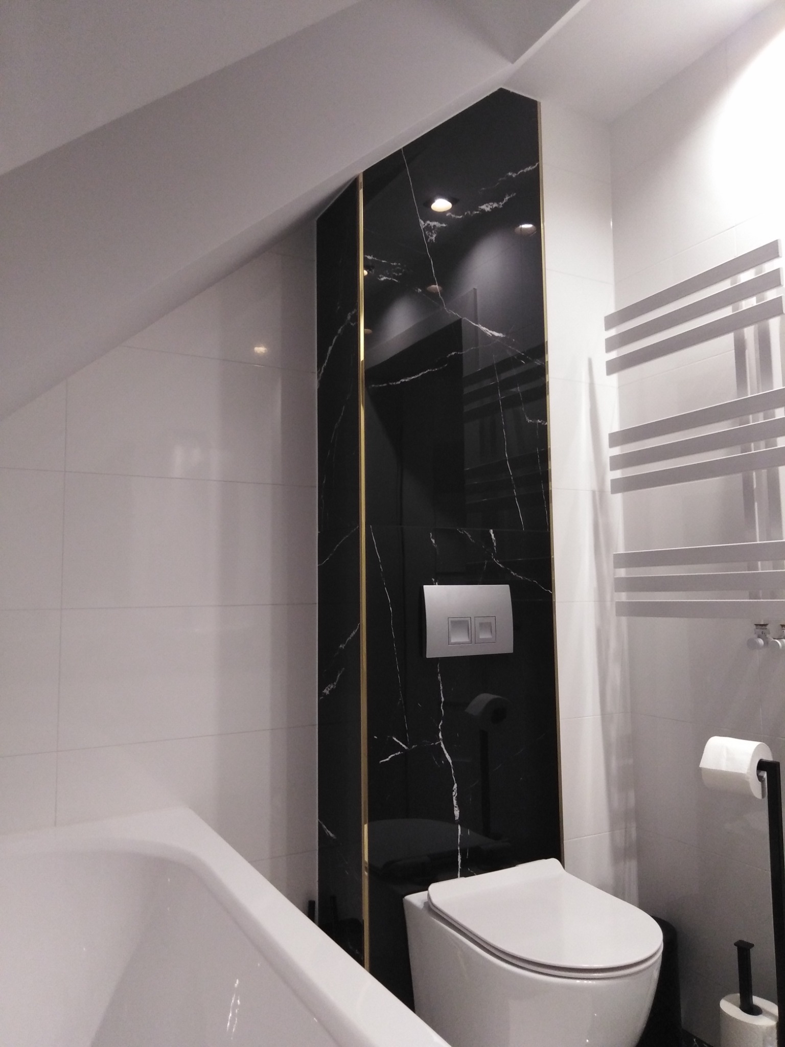 Nowoczesna łazienka w odcieniach bieli i czerni z akcentem złota, ukośna ściana, płytki imitujące marmur, podwieszana toaleta, grzejnik drabinkowy.