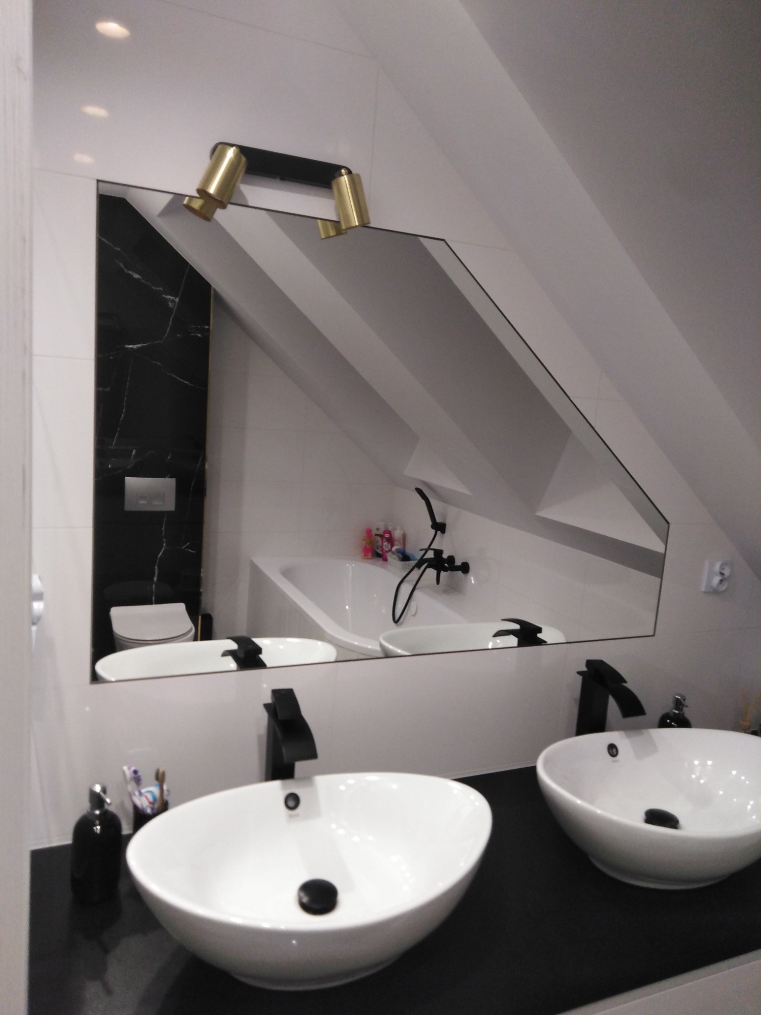 Nowoczesna łazienka na poddaszu z dwoma białymi umywalkami nablatowymi, czarnymi bateriami i blatem, lustrem odbijającym wannę i toaletę oraz złotymi lampkami nad lustrem.
