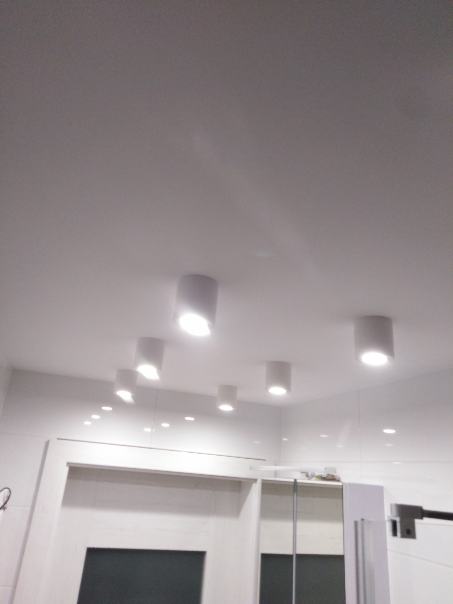 Biała łazienka z białymi, punktowymi lampami sufitowymi, odbijającymi się w błyszczących płytkach ściennych, widoczne fragmenty szafek i drzwi.