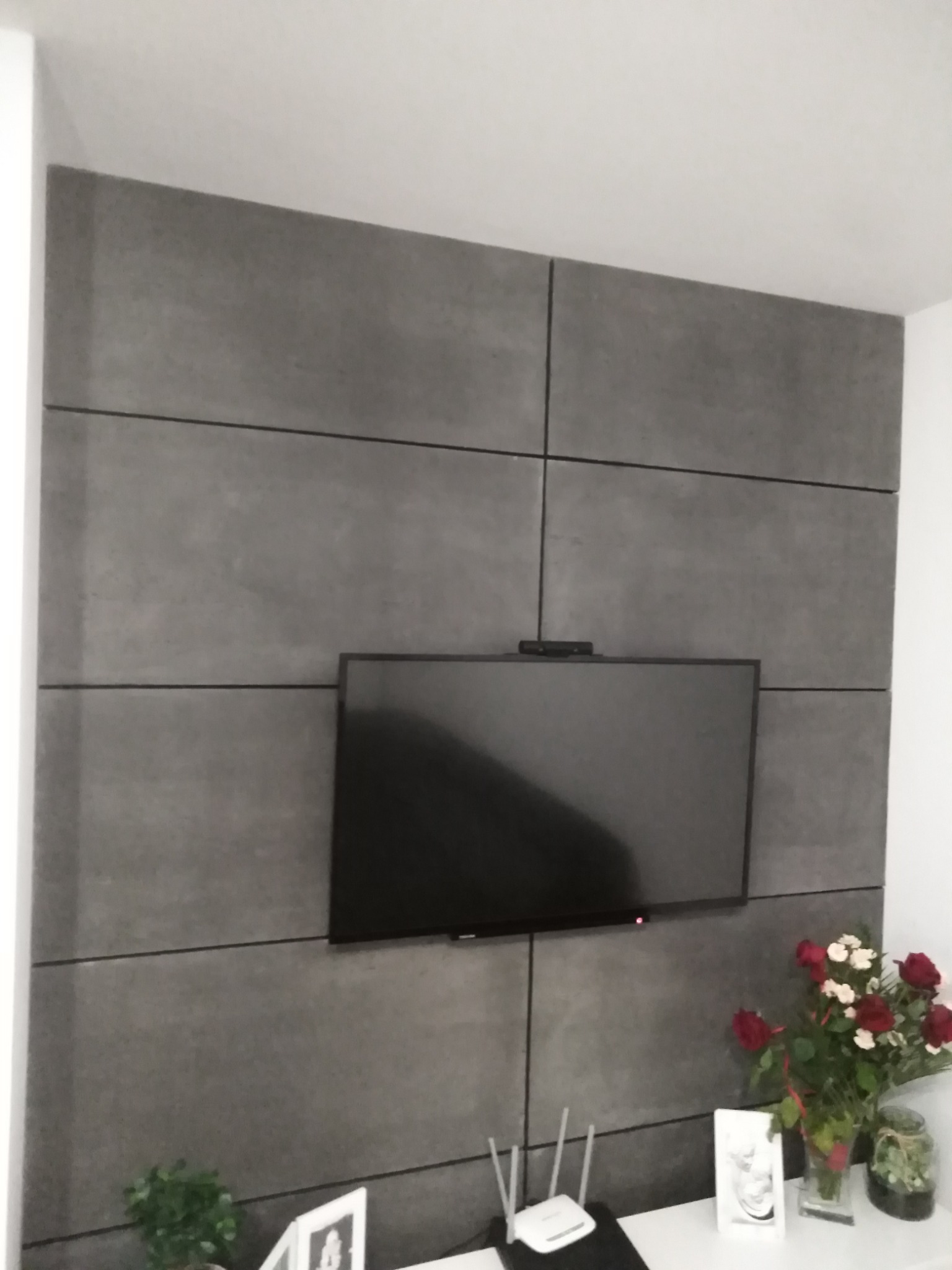 Telewizor zawieszony na ścianie wyłożonej panelami imitującymi beton, dekoracja kwiatowa i router na białej szafce RTV.