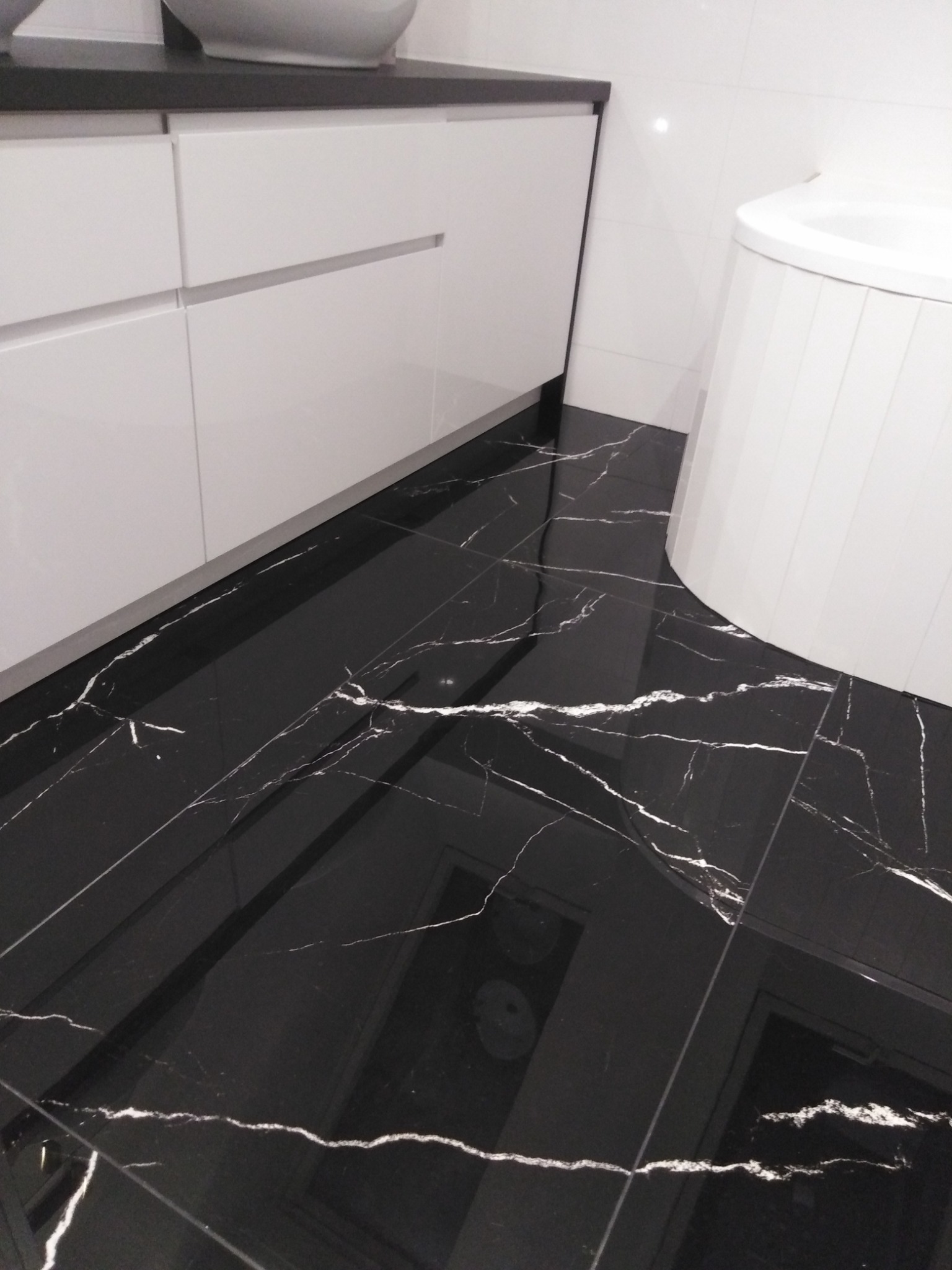 Błyszcząca, czarna podłoga z marmurkowym wzorem w łazience, widoczna dolna część białej szafki pod umywalkę i fragment wanny.