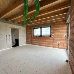 TERMA SYSTEM Patryk Kowalski - Wnętrze w stanie surowym z widoczną instalacją rekuperacji w postaci zielonych rur pod sufitem, przygotowaną podłogą z izolacją oraz instalacją elektryczną w ścianach z cegły.
