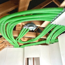 TERMA SYSTEM Patryk Kowalski - Zielone, karbowane rury wentylacyjne pod sufitem z drewnianą konstrukcją, widoczny rozdzielacz i fragment ściany w stanie surowym.