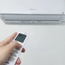 Biały klimatyzator Hisense na ścianie, sterowany pilotem; dłoń naciska przycisk regulacji prędkości wentylatora.