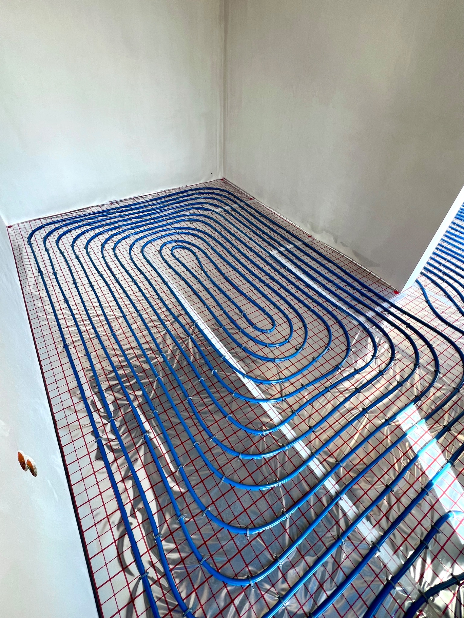 Niebieskie rury ogrzewania podłogowego ułożone w pętle na folii z siatką w surowym pomieszczeniu.