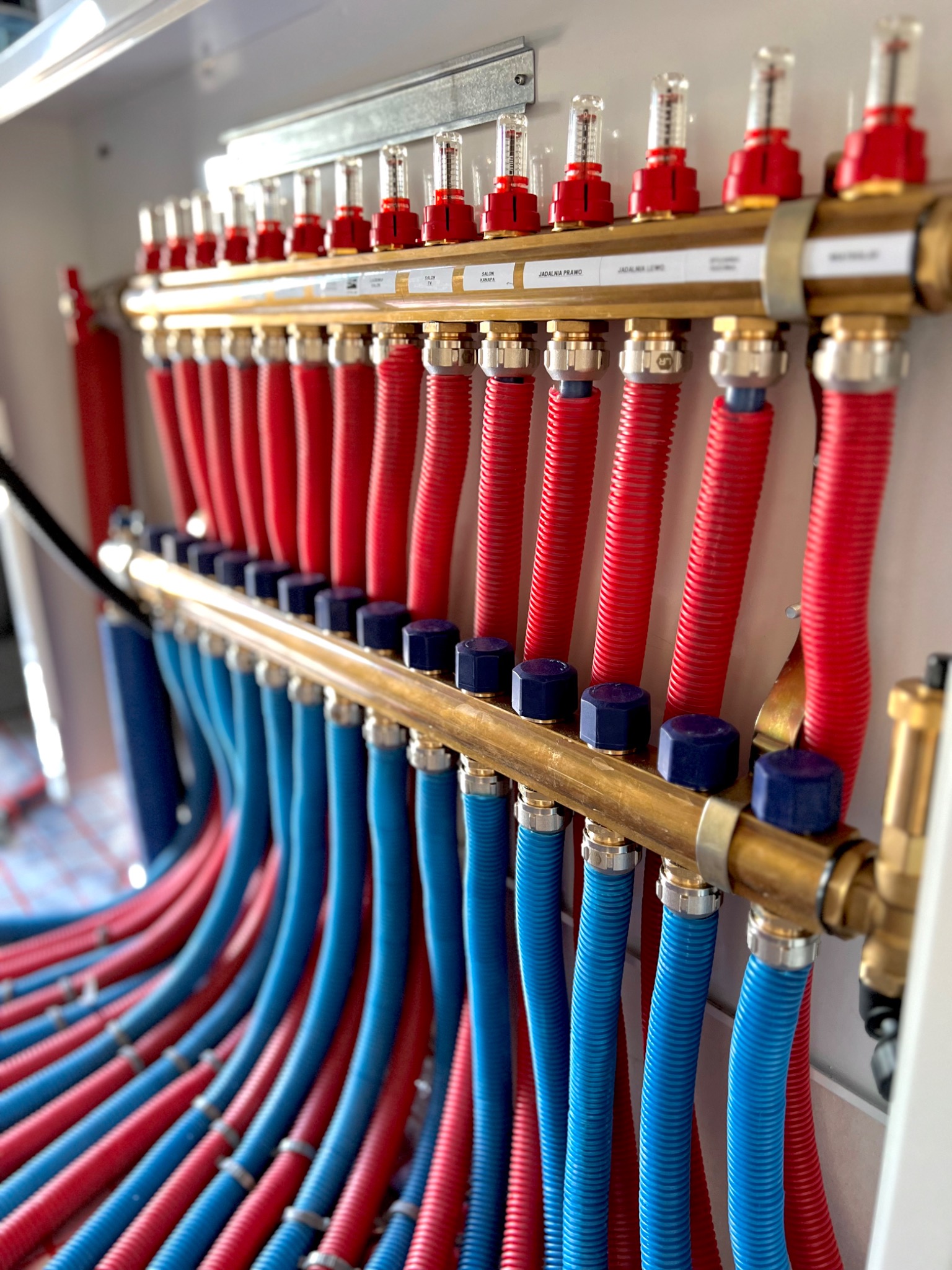 Rozdzielacz ogrzewania podłogowego z czerwonymi i niebieskimi rurami, etykiety z nazwami pomieszczeń nad górną belką, widok z dołu pod kątem.