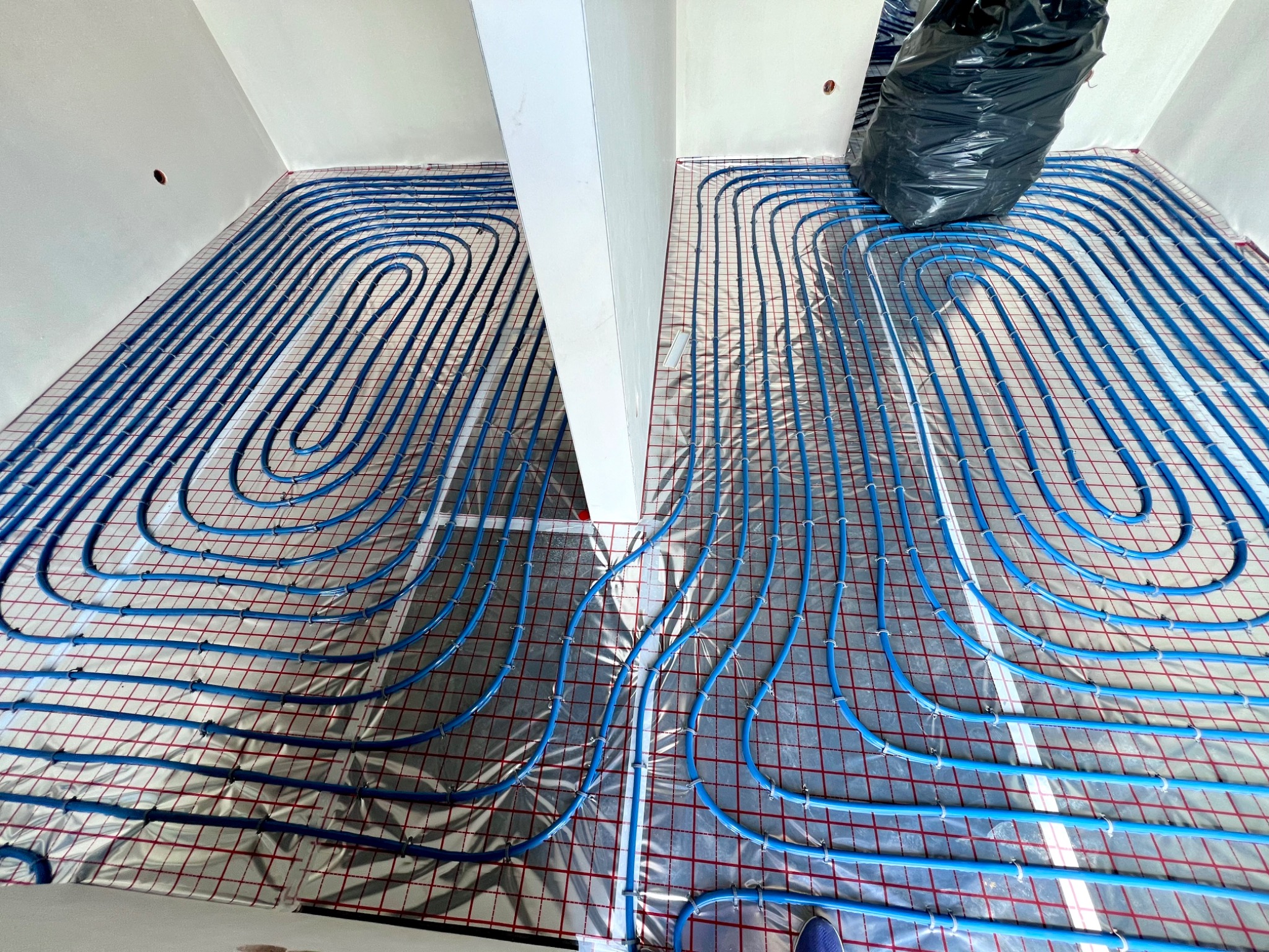 Niebieskie rury ogrzewania podłogowego rozłożone na folii z siatką w pomieszczeniu w Sadowicach, widok z góry, czarny worek na śmieci w rogu.