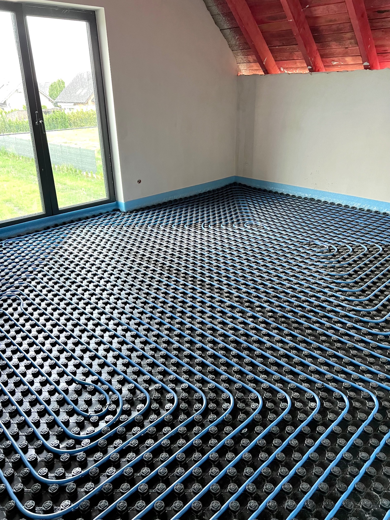 Niedokończona instalacja ogrzewania podłogowego w pomieszczeniu z oknem i odsłoniętą konstrukcją dachu. Równoległe rzędy niebieskich rurek ułożone na czarnej macie z wypustkami, zabezpieczone...