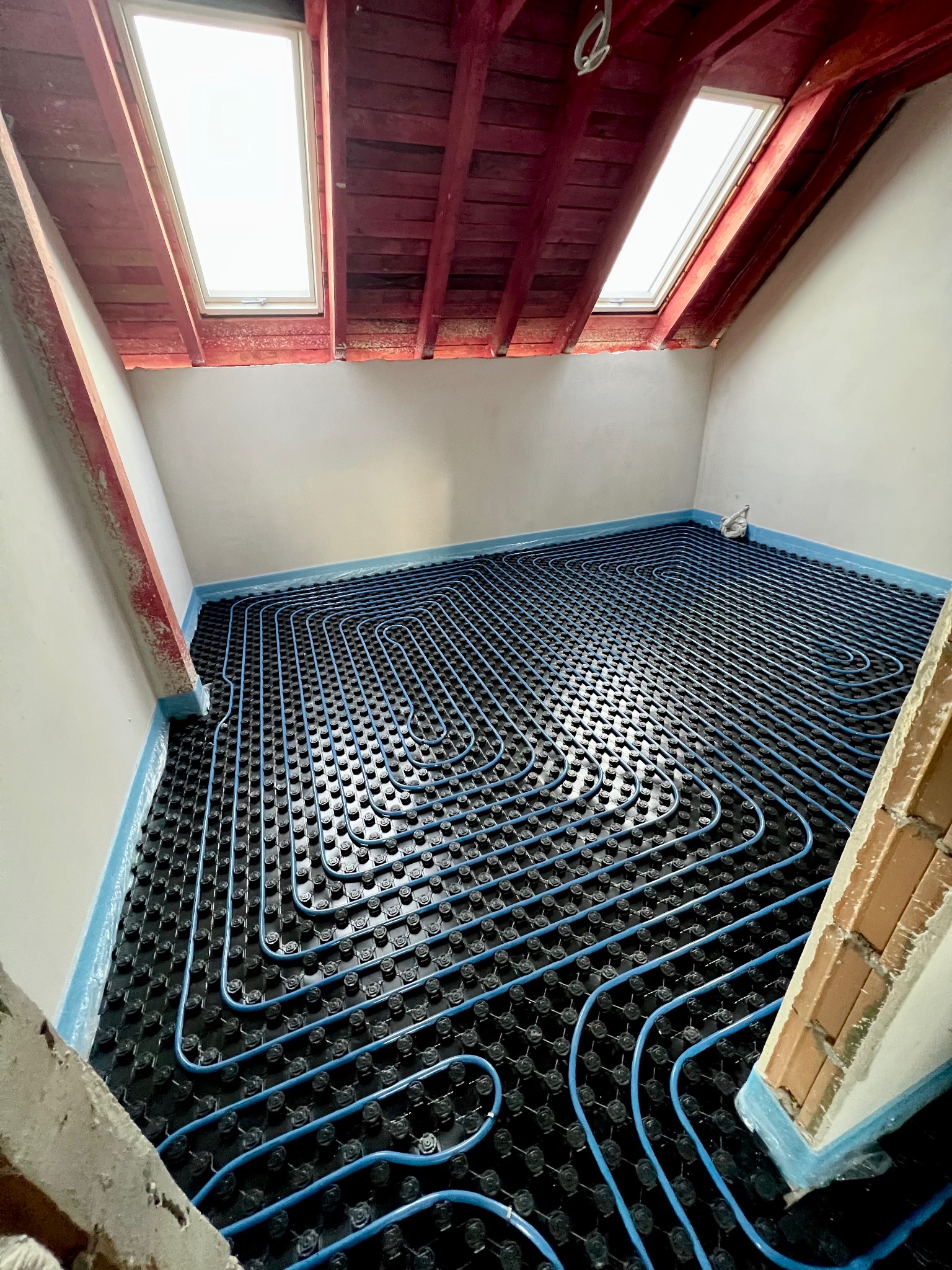 Niebieskie rury ogrzewania podłogowego ułożone spiralnie na czarnej macie w pokoju na poddaszu z oknami dachowymi, widoczne elementy konstrukcyjne budynku.