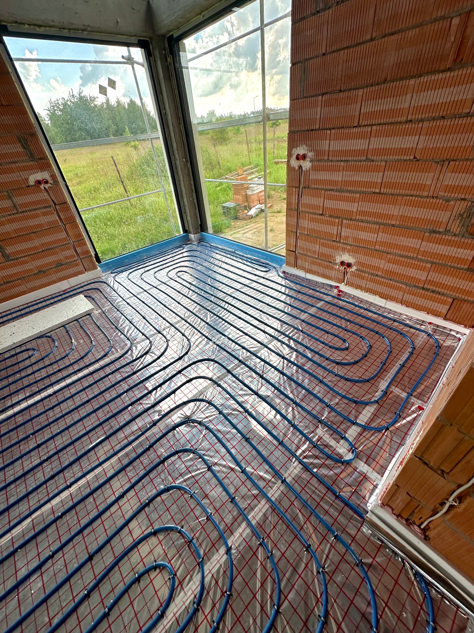 Niebieskie rury ogrzewania podłogowego, rozłożone na folii z czerwoną siatką, widok z góry na tle surowych cegieł i dużego okna z widokiem na zewnątrz.