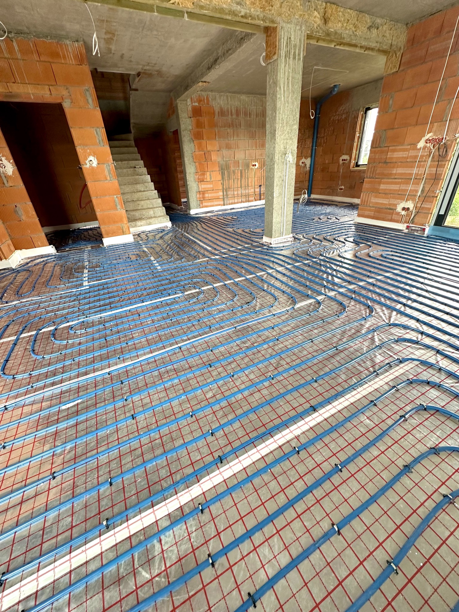 Niebieskie rury ogrzewania podłogowego ułożone na folii z czerwoną siatką w surowym stanie budynku, widoczne ceglane ściany i betonowe słupy.