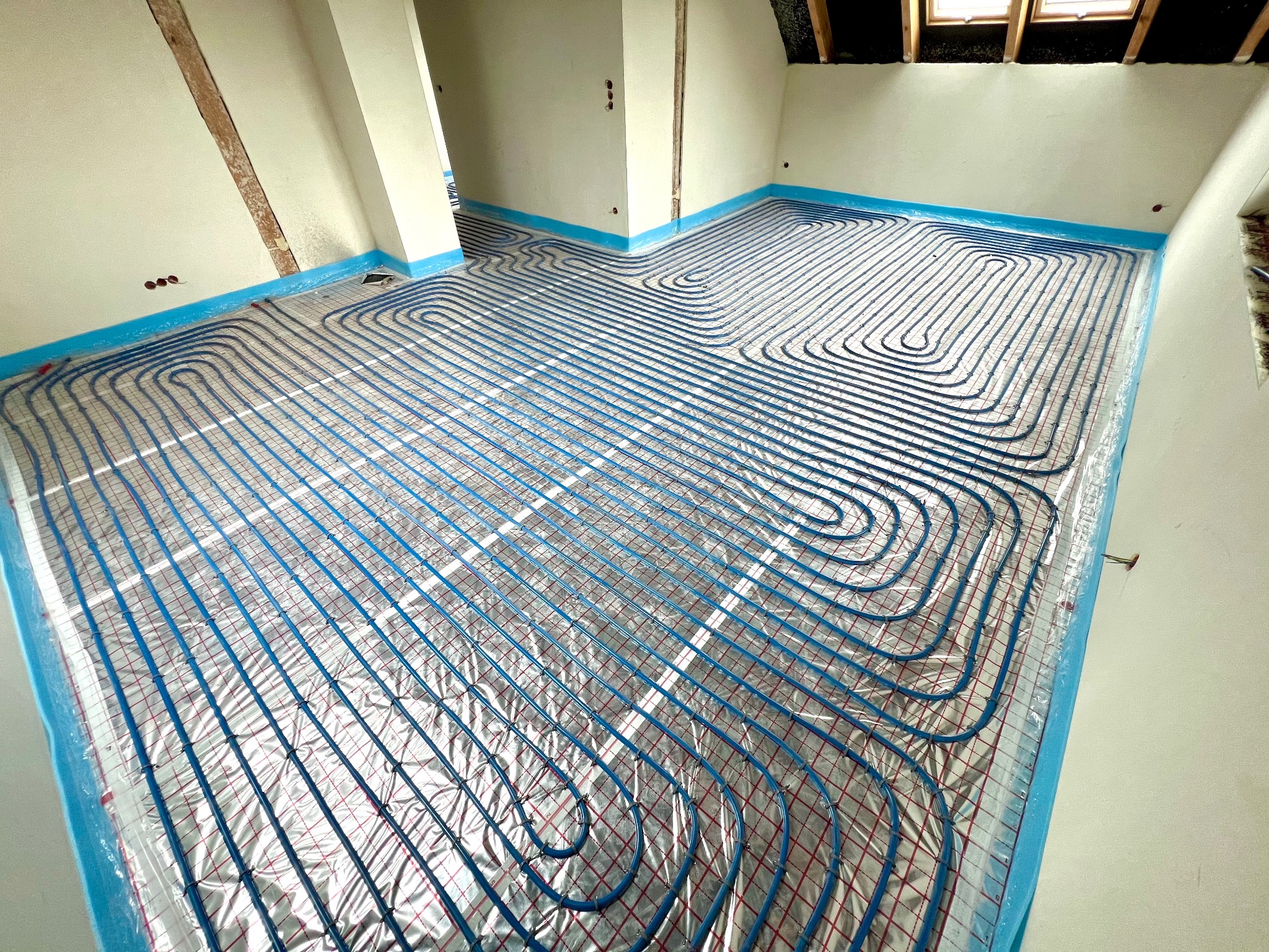 Niebieskie rury ogrzewania podłogowego ułożone na izolacji z folią aluminiową, zabezpieczone taśmą na ścianach w surowym wnętrzu poddasza.