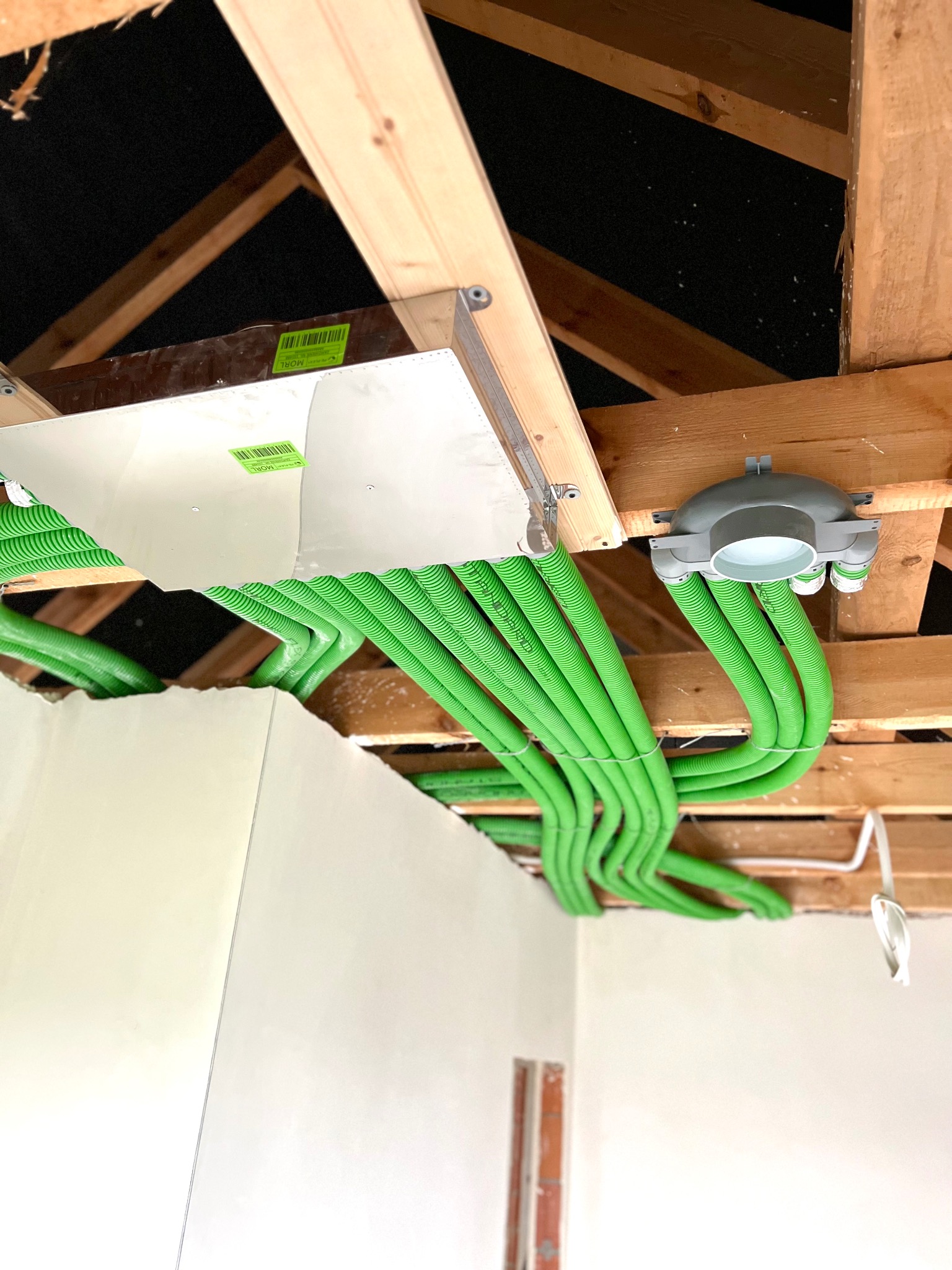 Widok z dołu na system rekuperacji z zielonymi przewodami wentylacyjnymi zainstalowany pod drewnianym stropem, widoczny fragment ściany w surowym stanie.