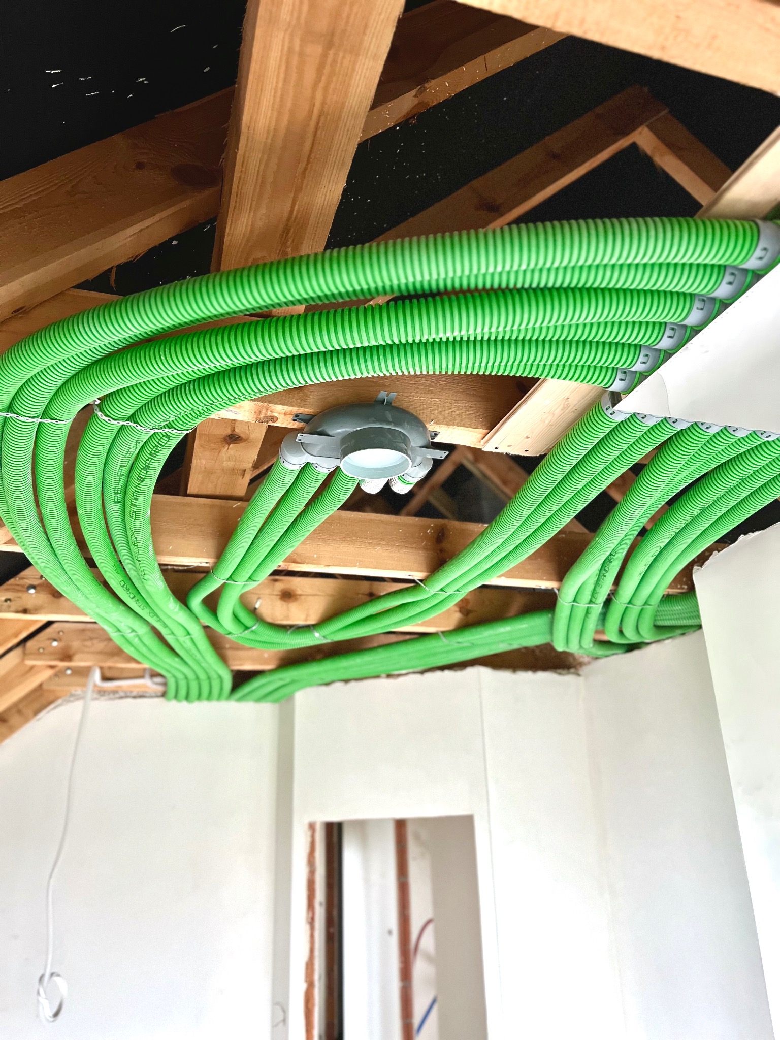 Zielone, karbowane rury wentylacyjne pod sufitem z drewnianą konstrukcją, widoczny rozdzielacz i fragment ściany w stanie surowym.