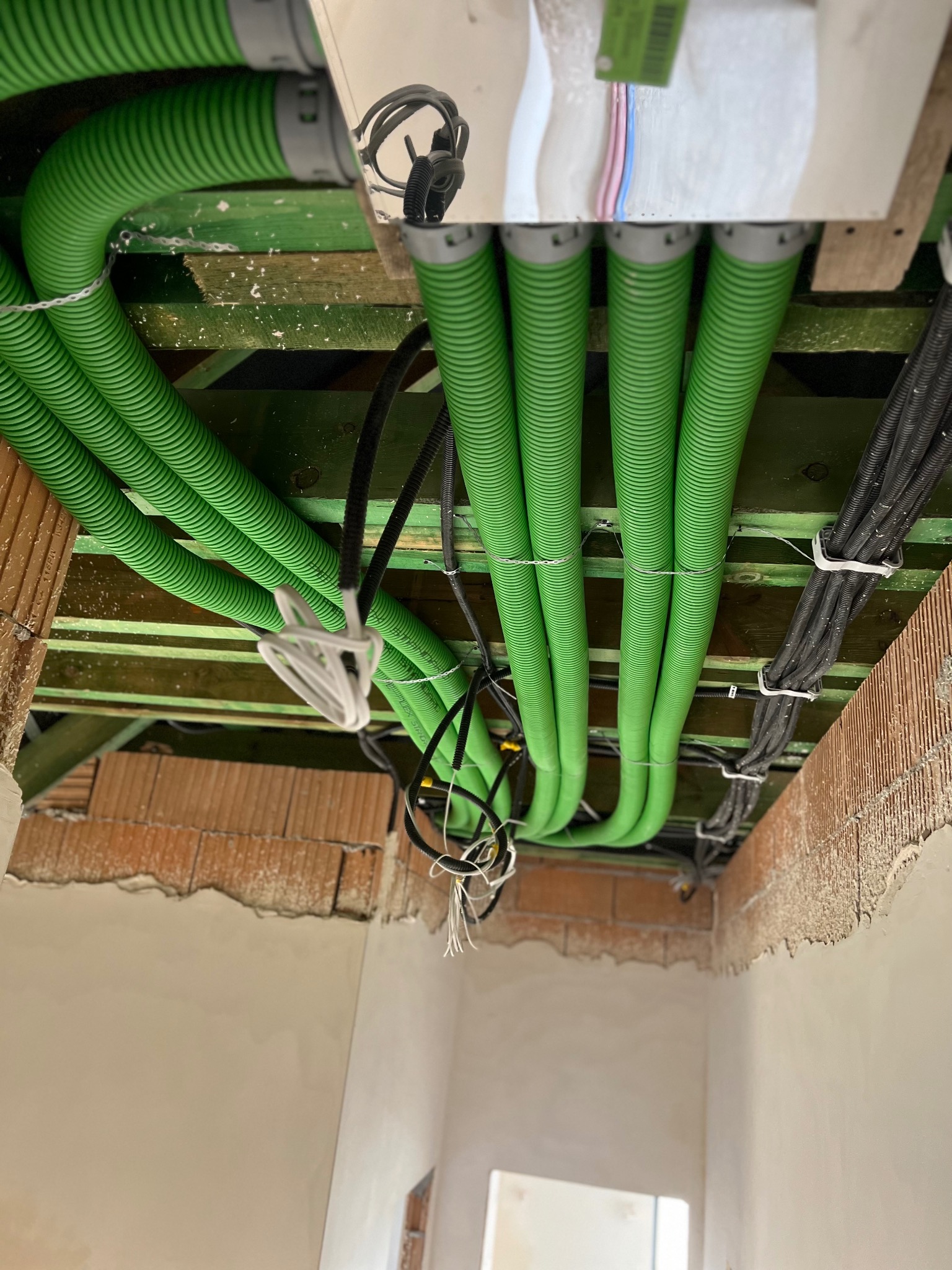 Zielone, karbowane rury wentylacyjne i czarne kable zamontowane pod sufitem w budynku w Sadowicach, widoczne elementy drewnianej konstrukcji i surowy mur.