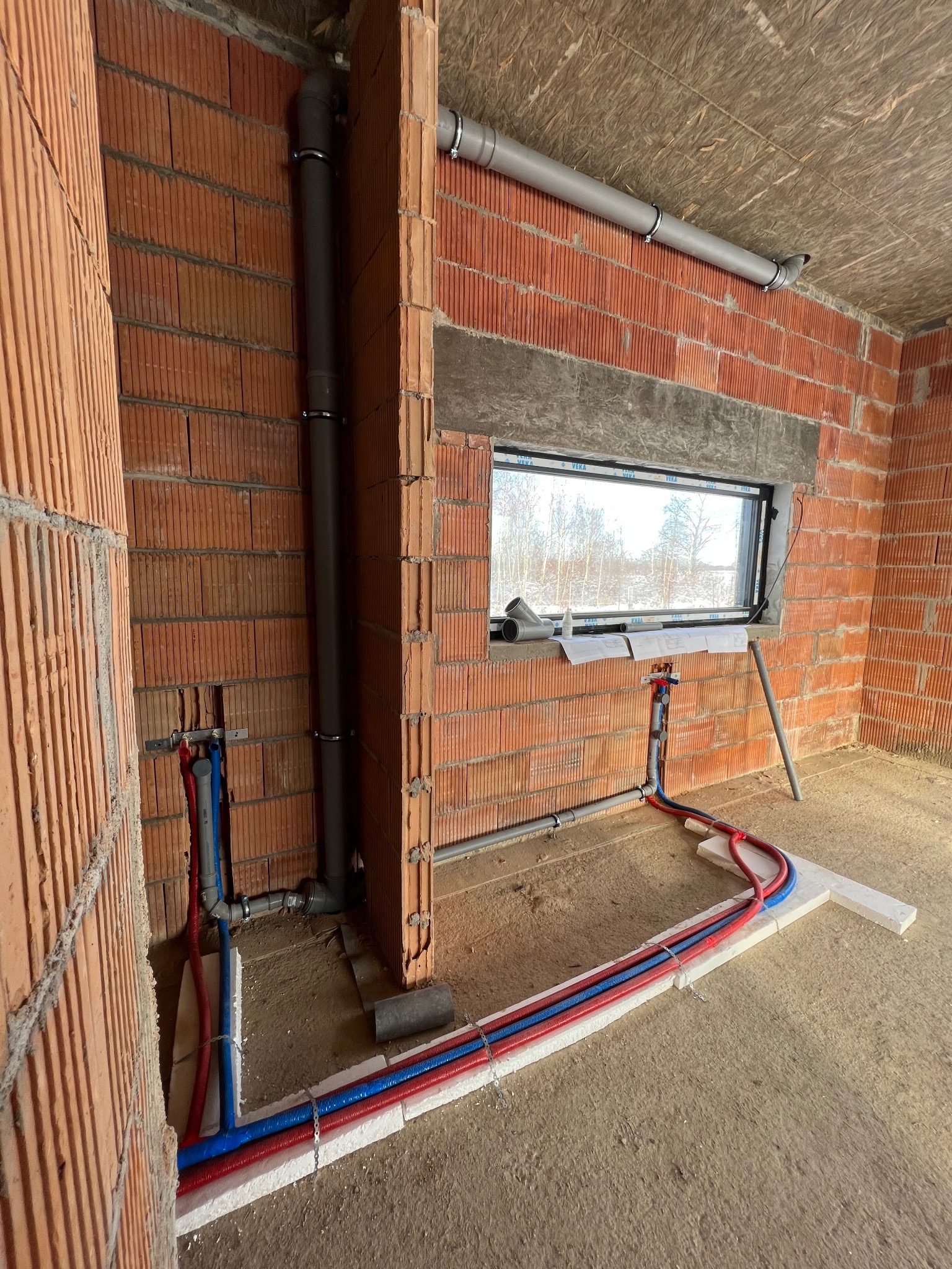 Instalacja hydrauliczna w surowym stanie: rury PEX w izolacji czerwonej i niebieskiej, pion kanalizacyjny, okno z widokiem na zimowy krajobraz, ściany z cegły dziurawki.