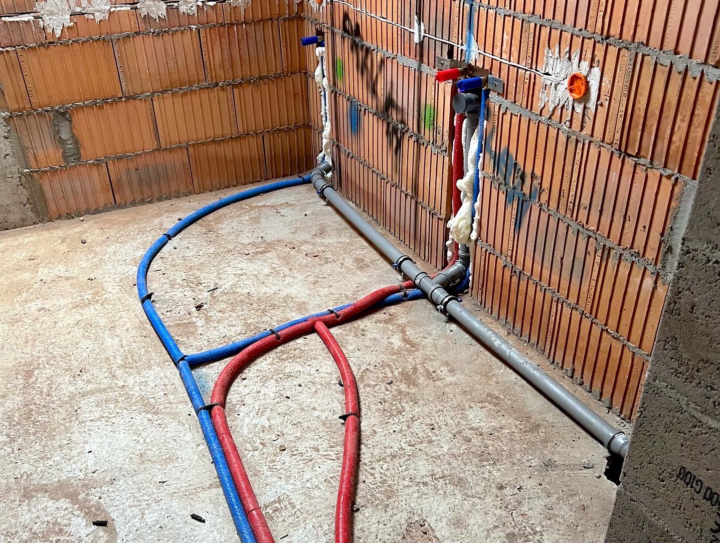 Wnętrze w trakcie prac hydraulicznych: czerwone i niebieskie rury PEX prowadzone po podłodze, szare rury PCV mocowane do ściany z czerwonej cegły, pianka montażowa uszczelniająca przejścia przez mur.