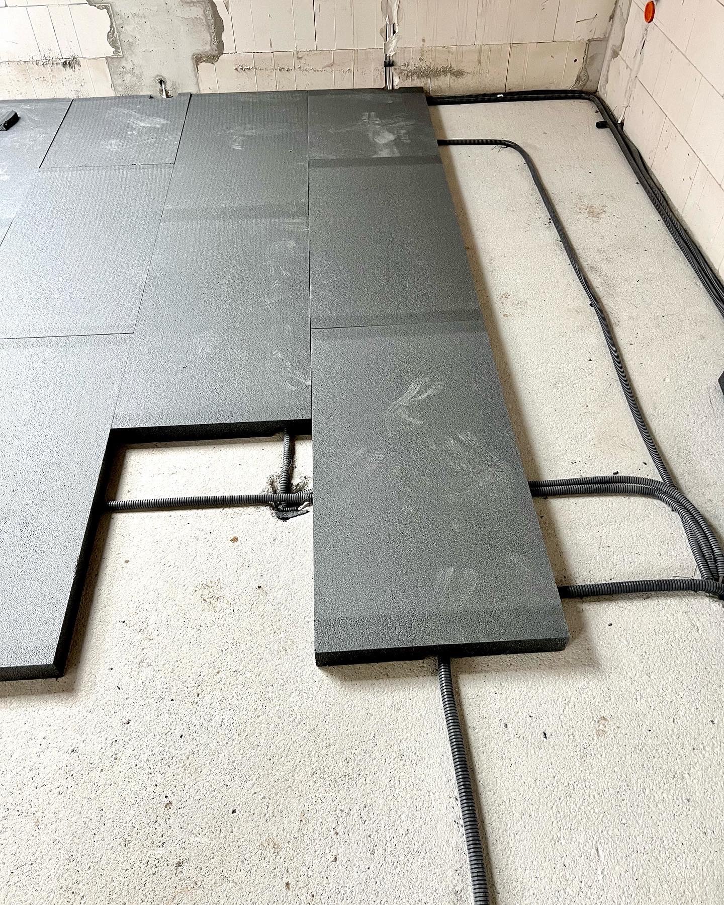 Przygotowanie podłoża pod ogrzewanie podłogowe: rozłożone płyty izolacyjne z widocznymi peszlami instalacyjnymi, surowy beton i ściany z betonu komórkowego w tle.