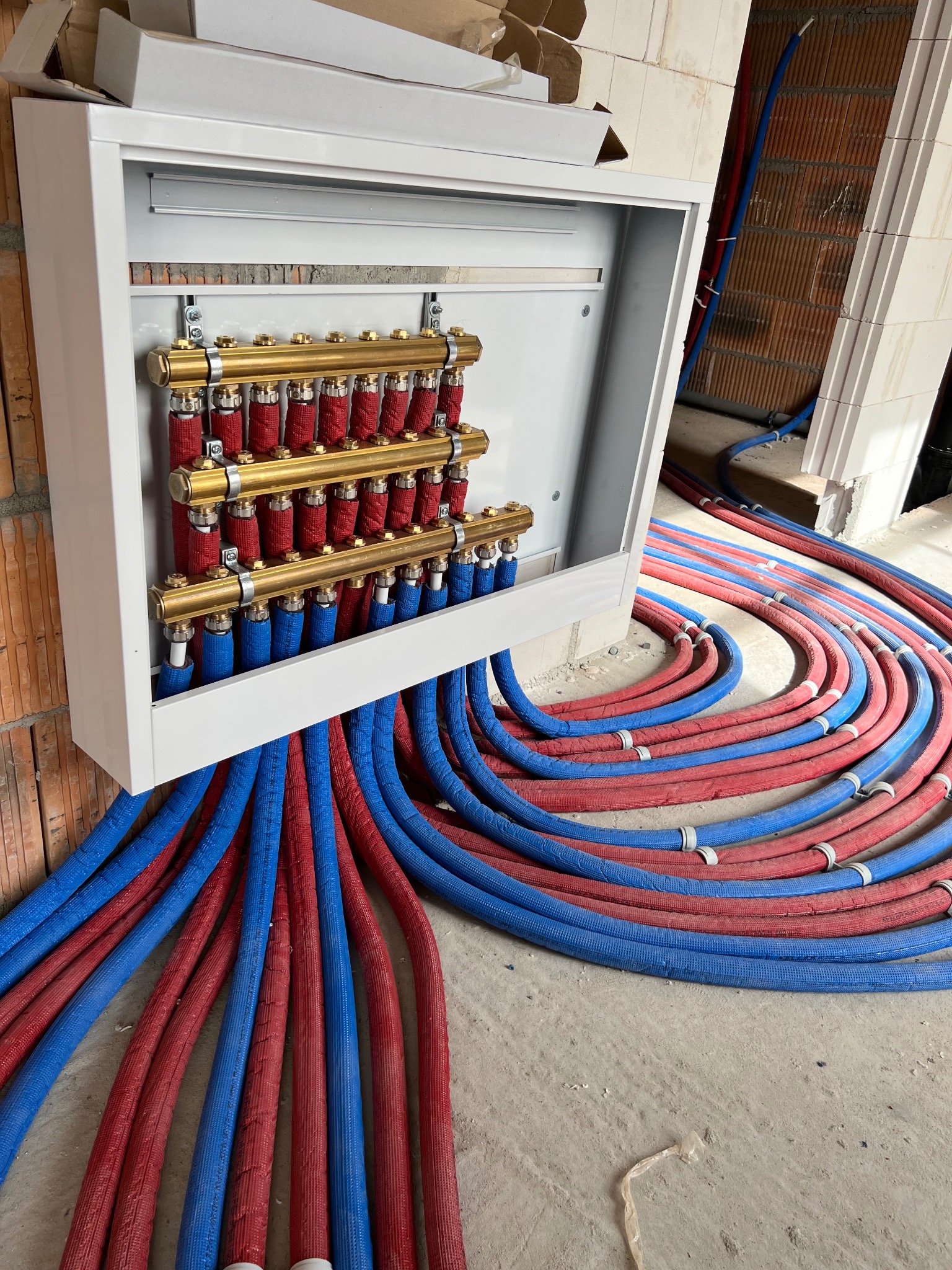 Rozdzielacz ogrzewania podłogowego w białej szafce, z licznymi rurami w kolorze czerwonym i niebieskim, ułożonymi na surowej posadzce w trakcie montażu.