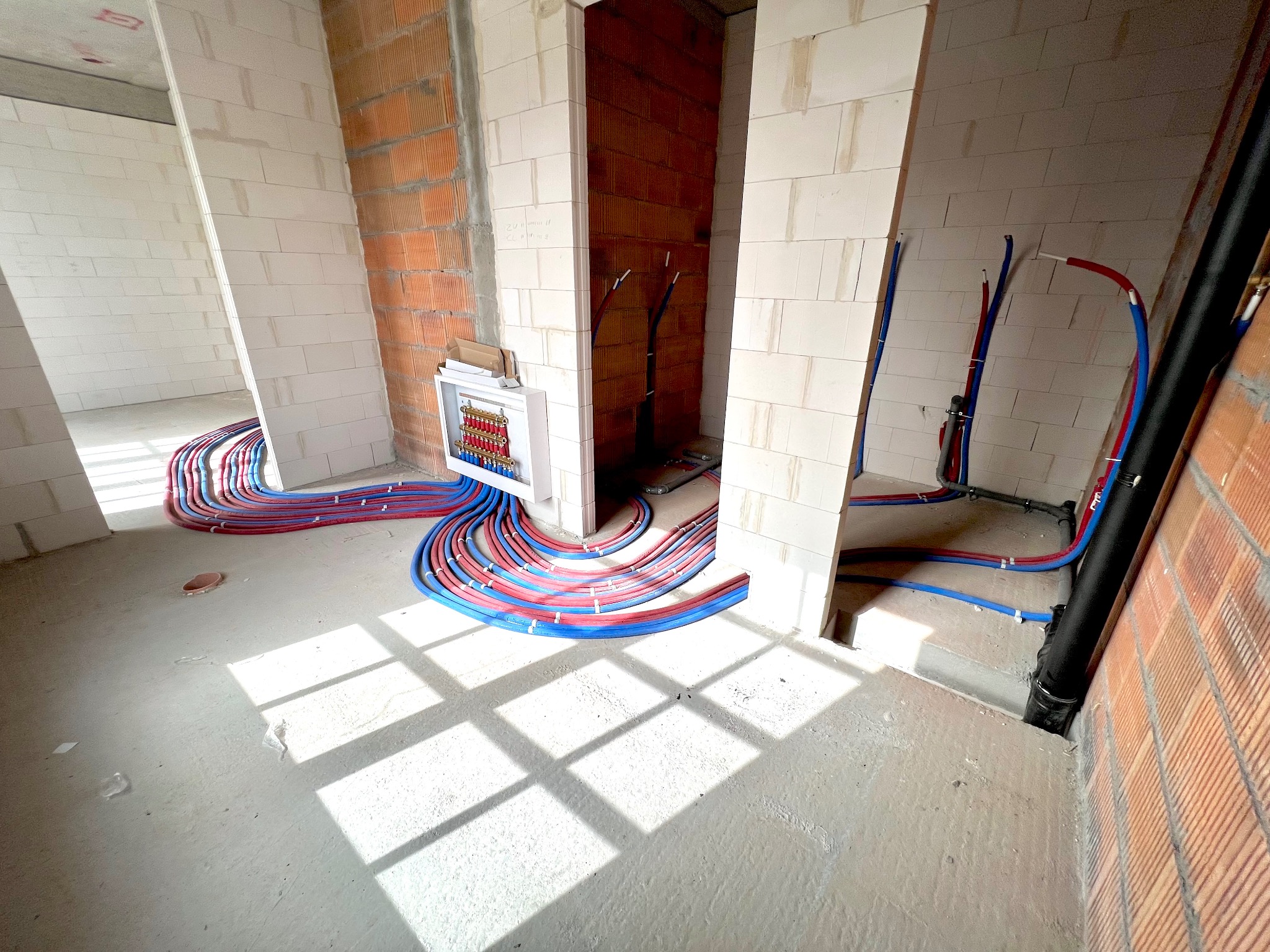 Wnętrze w stanie surowym z widoczną instalacją ogrzewania podłogowego: rozdzielacz w białej obudowie i pętle rur grzewczych w kolorach czerwonym i niebieskim, prowadzone wzdłuż ścian z bloczków...