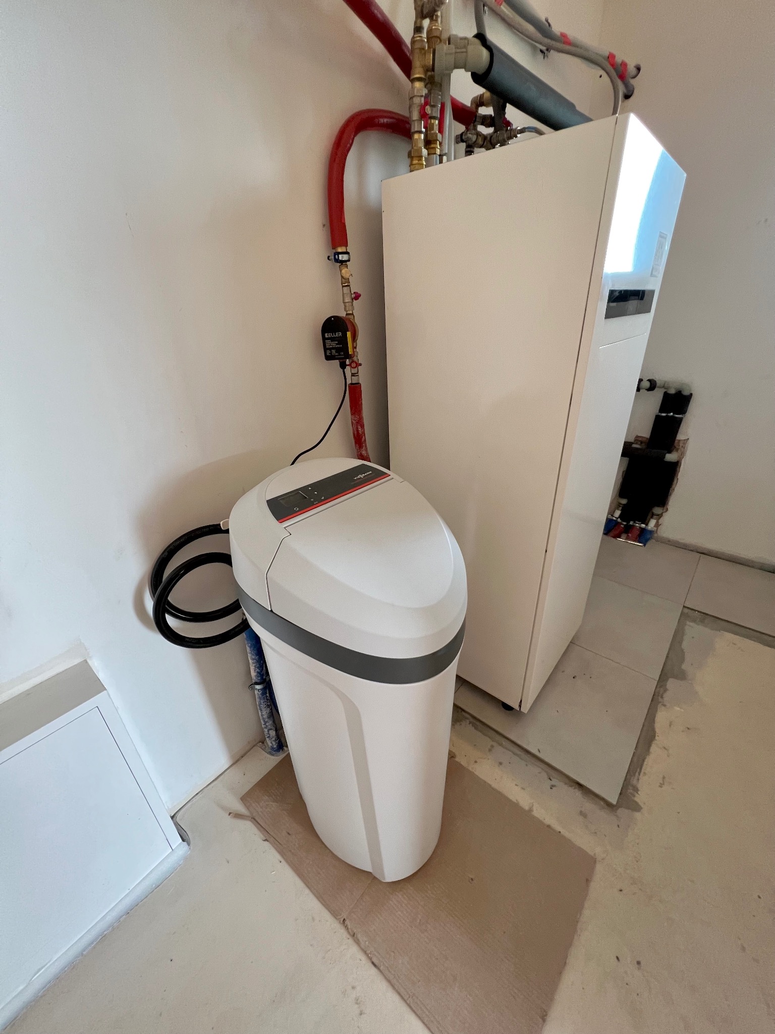 Zmiękczacz wody Viessmann obok białego pieca grzewczego z czerwonymi rurami w nowym pomieszczeniu z surową podłogą.