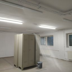 Mal-Nat Malowanie Natryskowe - Szare metalowe szafki w pustym pomieszczeniu z oknami i grzejnikami, widoczna puszka z farbą na podłodze, sufit z lampami i instalacją elektryczną.