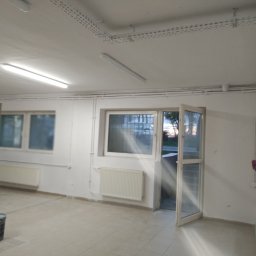 Mal-Nat Malowanie Natryskowe - Pomalowane na biało wnętrze pomieszczenia z grzejnikami pod oknami, otwartymi drzwiami balkonowymi i wiaderkiem z farbą w rogu, sugerujące świeżo zakończone prace malarskie.