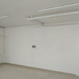 Mal-Nat Malowanie Natryskowe - Puste, świeżo pomalowane pomieszczenie z białymi ścianami, widoczne białe listwy kablowe pod sufitem, otwarte drzwi balkonowe i włącznik światła z czerwonymi i niebieskimi przyciskami.