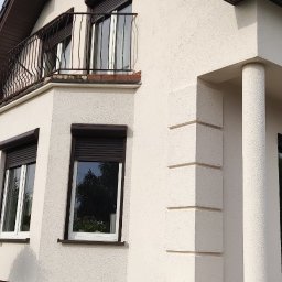 Mal-Nat Malowanie Natryskowe - Fragment elewacji domu z oknami z roletami zewnętrznymi, balkonem z metalową balustradą i schodami wejściowymi z kostki betonowej, wykończony w kolorze beżowym.