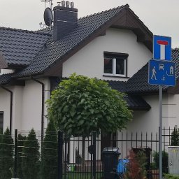 Mal-Nat Malowanie Natryskowe - Dom jednorodzinny z szarą dachówką, częściowo otoczony rusztowaniem, sugerujący prace remontowe lub malowanie elewacji. Widoczny fragment ogrodzenia i drzewo przed budynkiem.