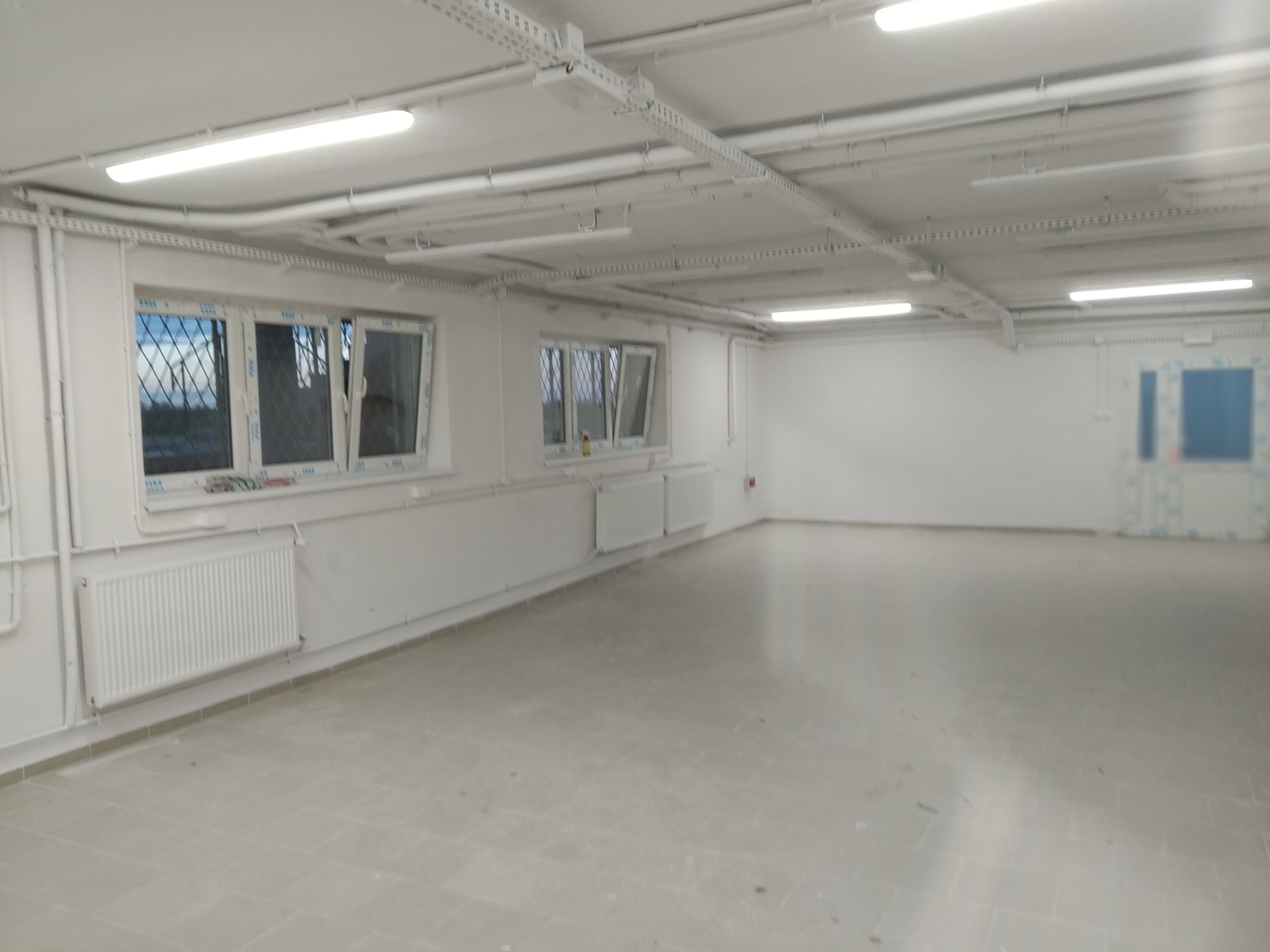Jasne, puste pomieszczenie z białymi ścianami, grzejnikami i oknami z kratami, oświetlone lampami sufitowymi i z szarą podłogą z płytek.