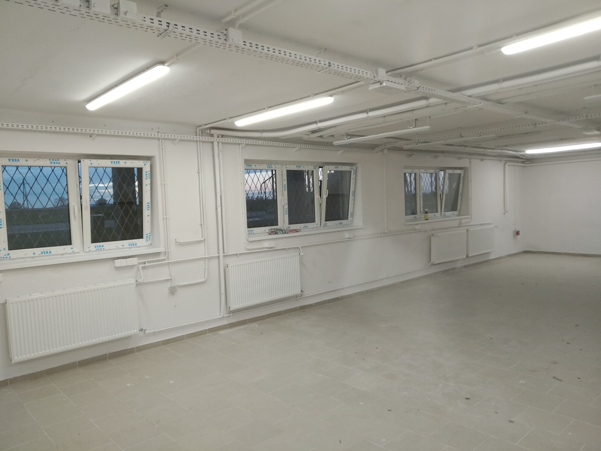 Wnętrze po remoncie: białe ściany, okna z charakterystycznymi szprosami, grzejniki i instalacje pod sufitem, jasne płytki na podłodze, widok z perspektywy szerokokątnej.