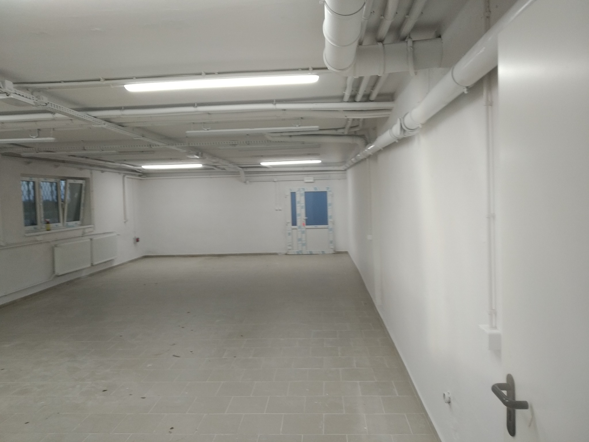Puste, wykończone pomieszczenie z szarą podłogą z płytek, białymi ścianami i sufitem z widocznymi rurami wentylacyjnymi oraz oświetleniem liniowym, widok na okno i drzwi w oddali.