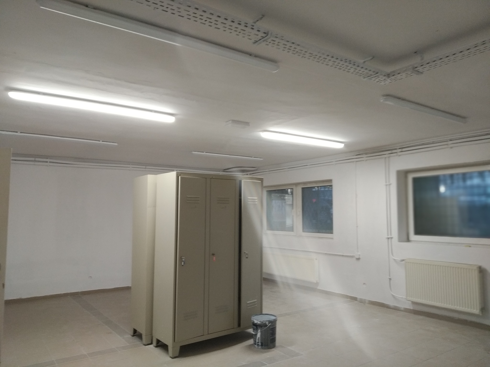 Szare metalowe szafki w pustym pomieszczeniu z oknami i grzejnikami, widoczna puszka z farbą na podłodze, sufit z lampami i instalacją elektryczną.