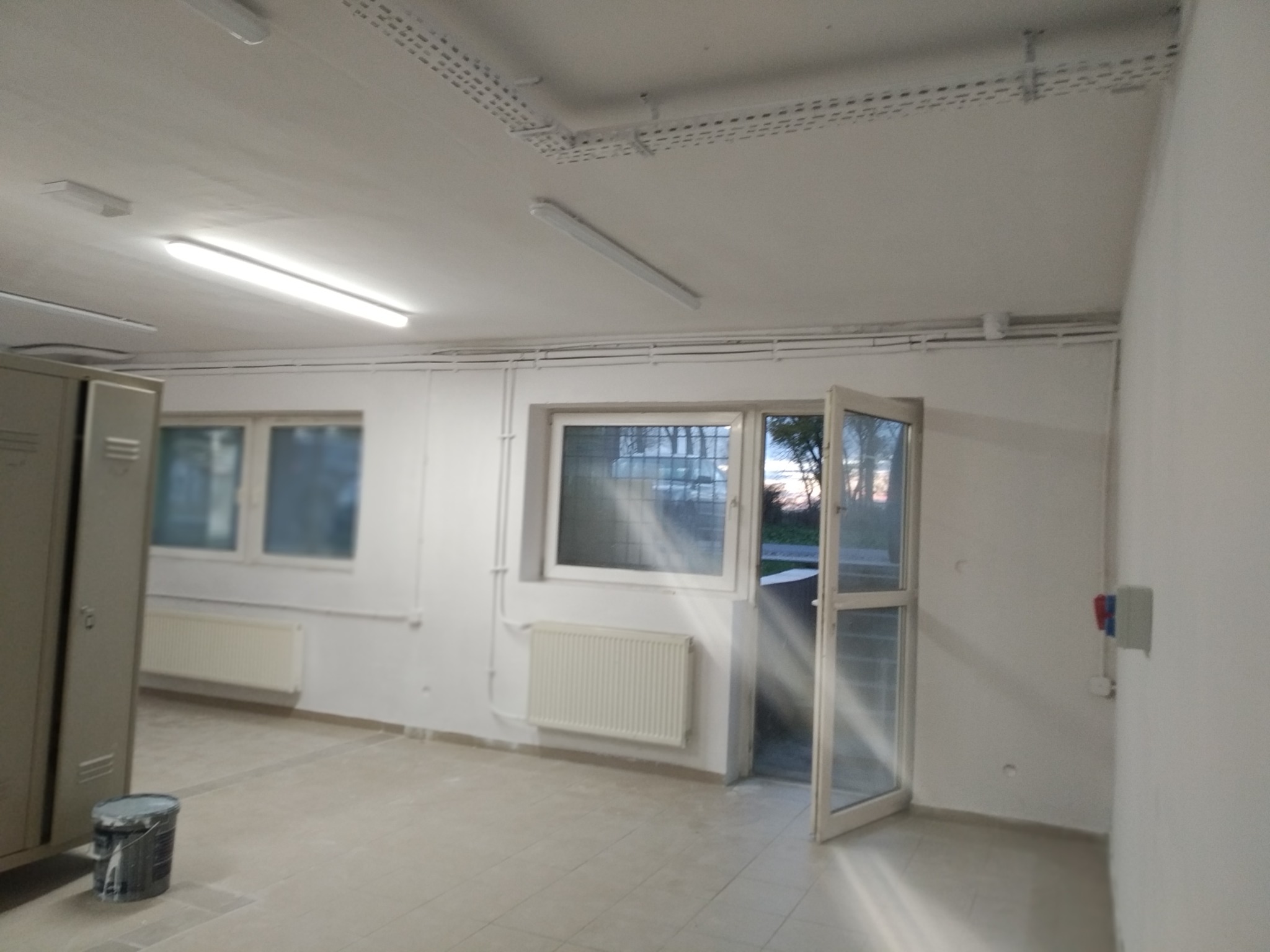 Pomalowane na biało wnętrze pomieszczenia z grzejnikami pod oknami, otwartymi drzwiami balkonowymi i wiaderkiem z farbą w rogu, sugerujące świeżo zakończone prace malarskie.
