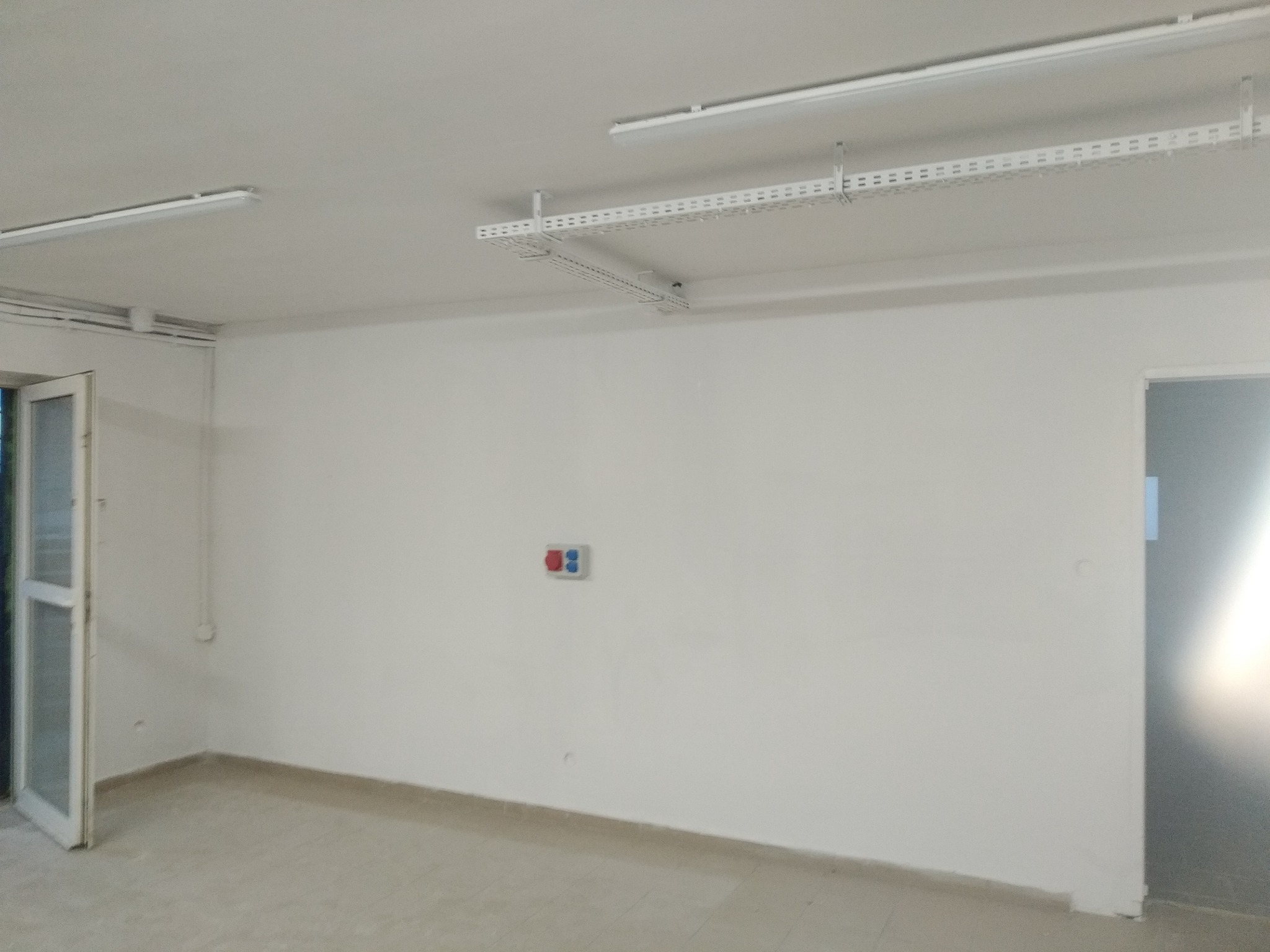 Puste, świeżo pomalowane pomieszczenie z białymi ścianami, widoczne białe listwy kablowe pod sufitem, otwarte drzwi balkonowe i włącznik światła z czerwonymi i niebieskimi przyciskami.