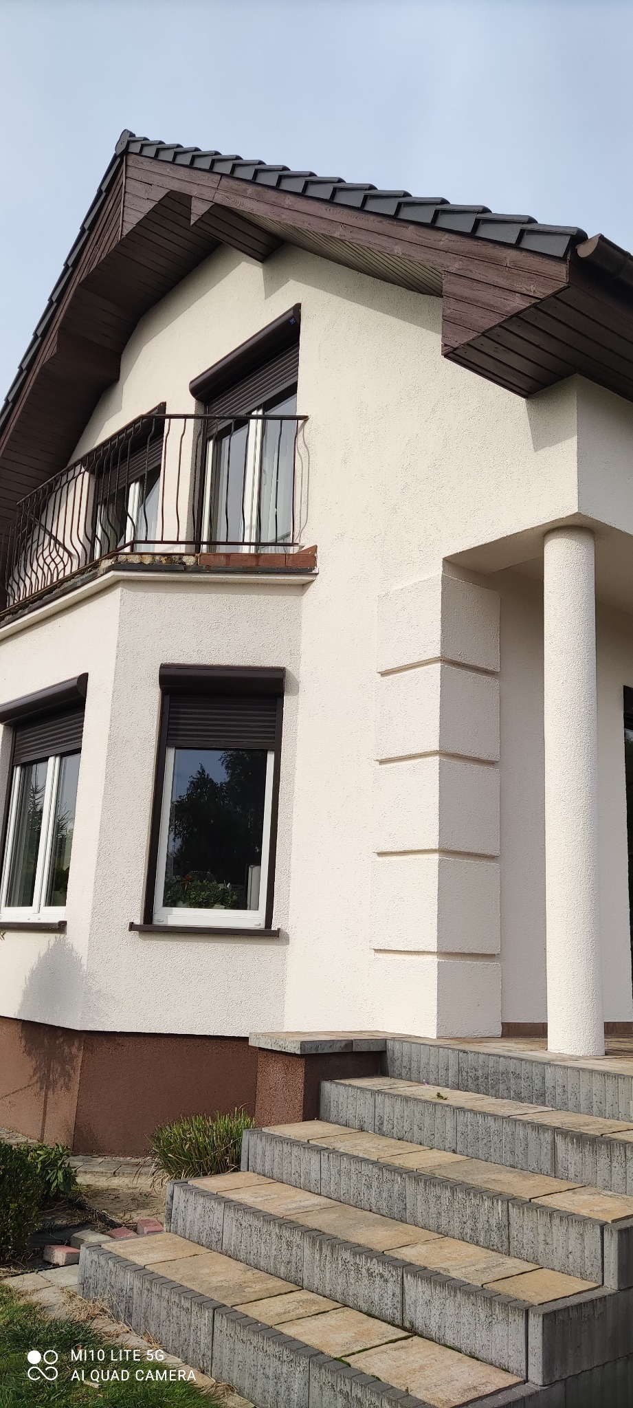 Fragment elewacji domu z oknami z roletami zewnętrznymi, balkonem z metalową balustradą i schodami wejściowymi z kostki betonowej, wykończony w kolorze beżowym.