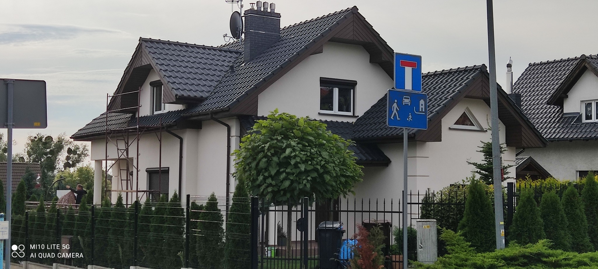 Dom jednorodzinny z szarą dachówką, częściowo otoczony rusztowaniem, sugerujący prace remontowe lub malowanie elewacji. Widoczny fragment ogrodzenia i drzewo przed budynkiem.