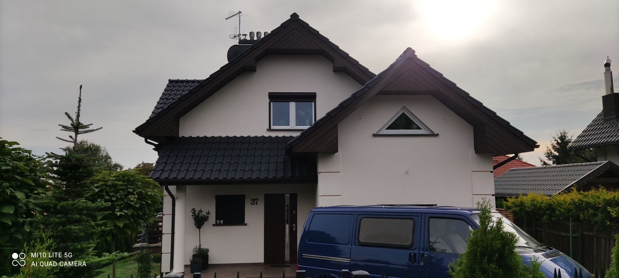 Dwukondygnacyjny dom jednorodzinny z białymi ścianami, ciemnobrązowym dachem i stolarką okienną, z numerem 37 nad drzwiami, częściowo zasłonięty przez niebieskiego vana i zieleń ogrodu.