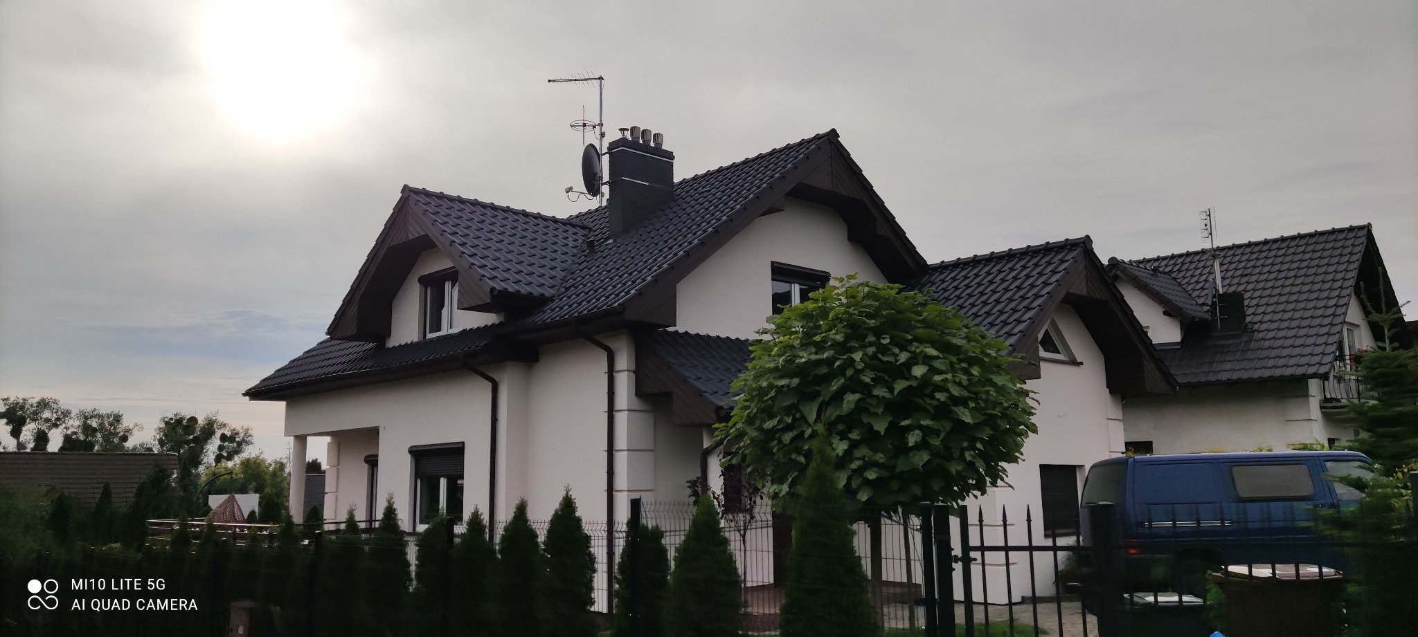 Dwupiętrowy dom z ciemnym dachem i jasną elewacją, widoczny zza ogrodzenia z żywopłotu i ozdobnego drzewa, z zaparkowanym niebieskim vanem w tle, w pochmurny dzień.