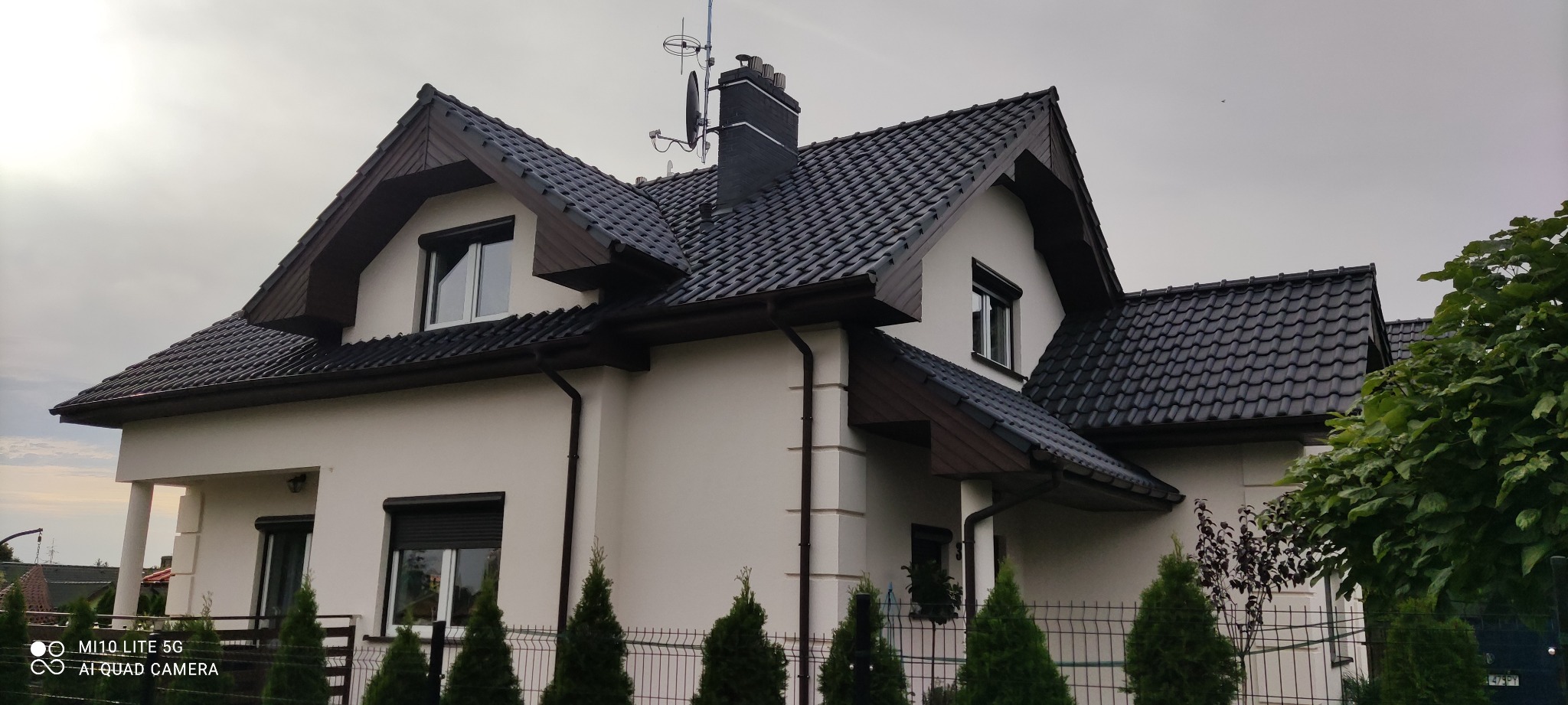 Dom jednorodzinny z kremową elewacją, ciemną dachówką i brązowymi elementami wykończeniowymi, sfotografowany z poziomu gruntu, ukazujący front budynku i część ogrodu z iglakami.