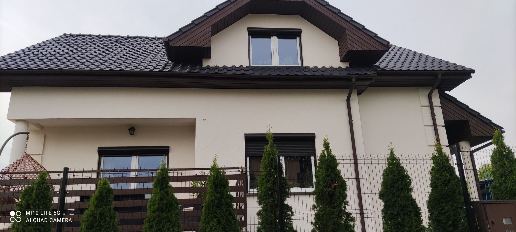 Elewacja domu jednorodzinnego z oknami z roletami zewnętrznymi, ogrodzona ozdobnymi tujami w Poznaniu.