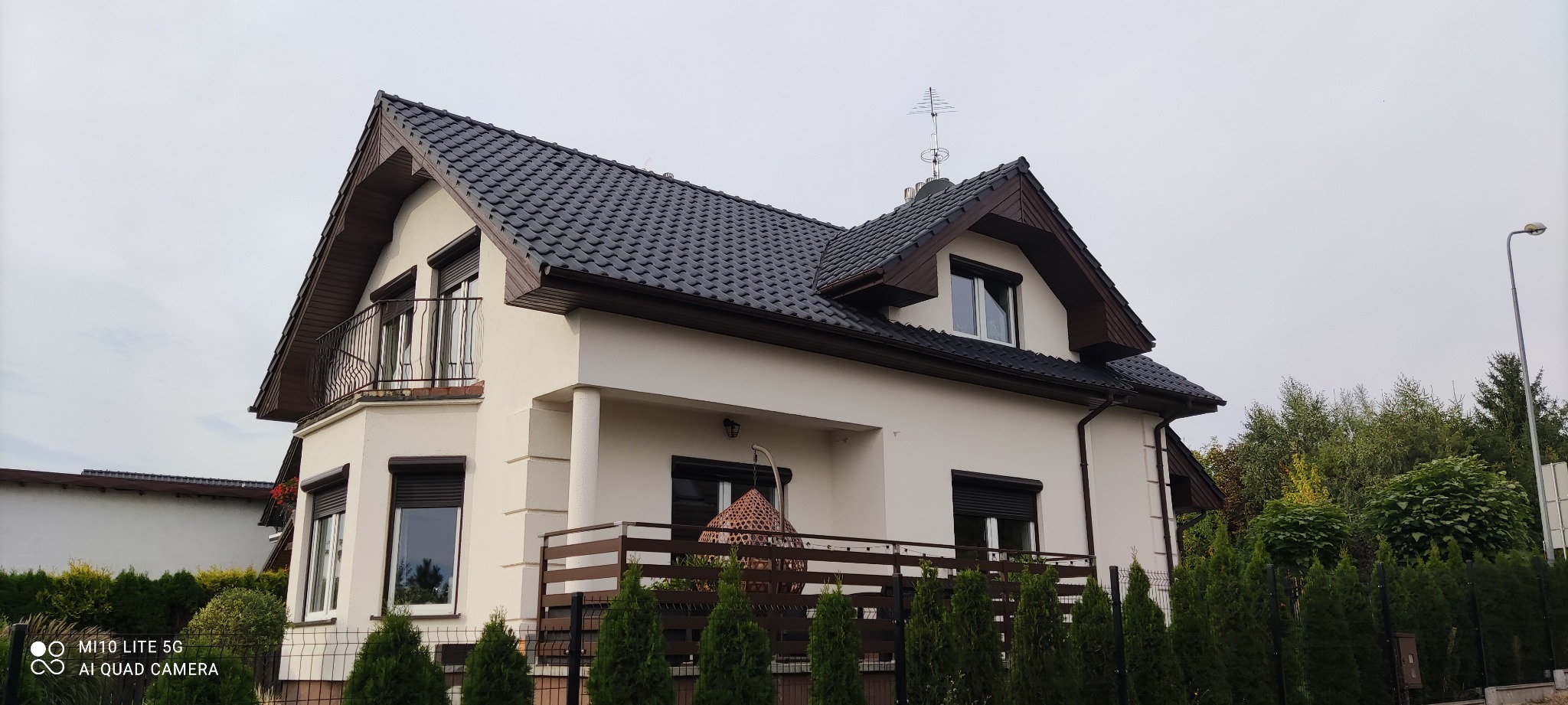 Dwupiętrowy dom z beżową elewacją, ciemnym dachem z dachówki, balkonem z kutą balustradą i drewnianym tarasem z wiszącym fotelem rattanowym, otoczony tujami.