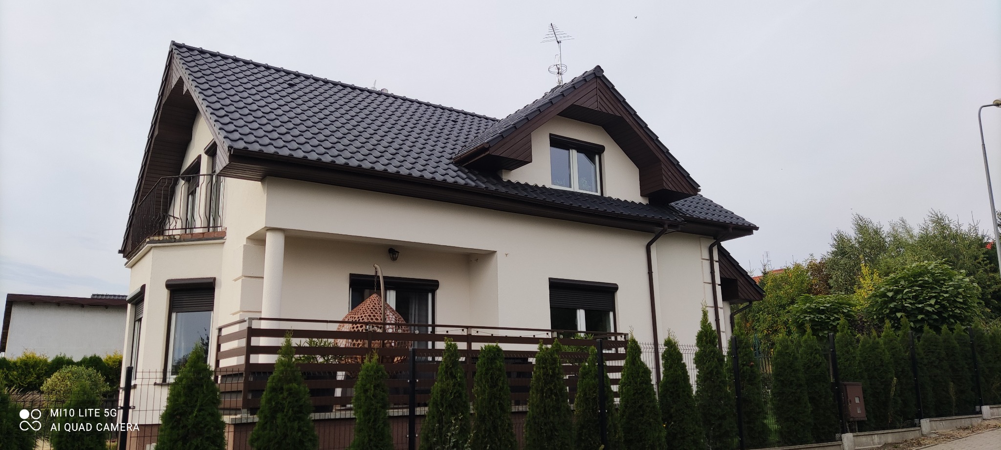 Dwupiętrowy dom z balkonem i ciemną dachówką, widok z zewnątrz, z ozdobnym fotelem wiszącym na balkonie i rzędem małych drzewek iglastych wzdłuż ogrodzenia.