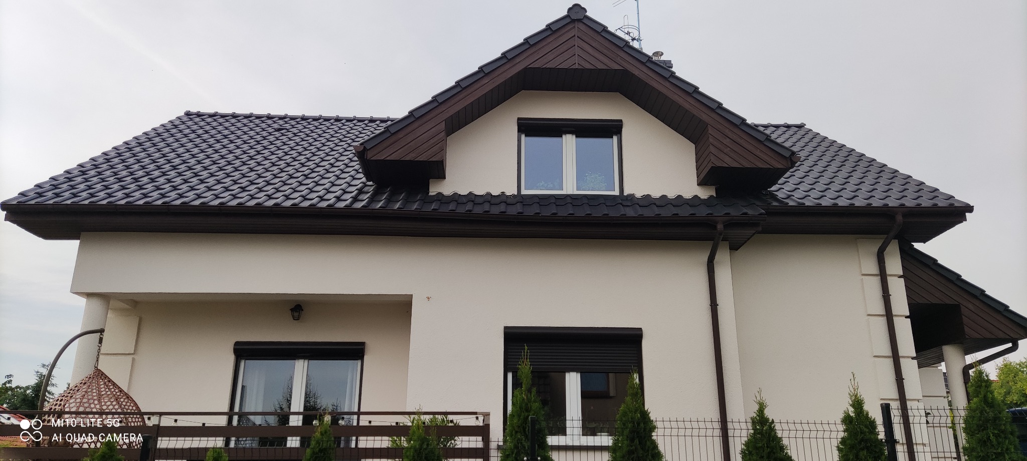 Elewacja domu jednorodzinnego w kolorze kremowym z ciemnobrązową dachówką, rynnami i podbitką, widoczne okna z roletami zewnętrznymi i zadbane tuje wzdłuż ogrodzenia.