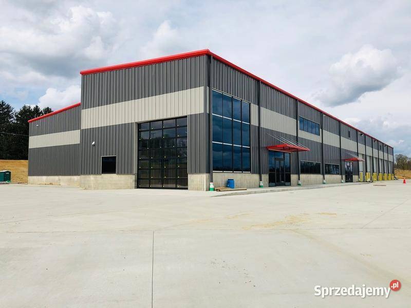 Nowoczesny budynek przemysłowy z szarej blachy trapezowej z czerwonymi akcentami na dachu i nad wejściem, z dużymi oknami i bramą garażową.