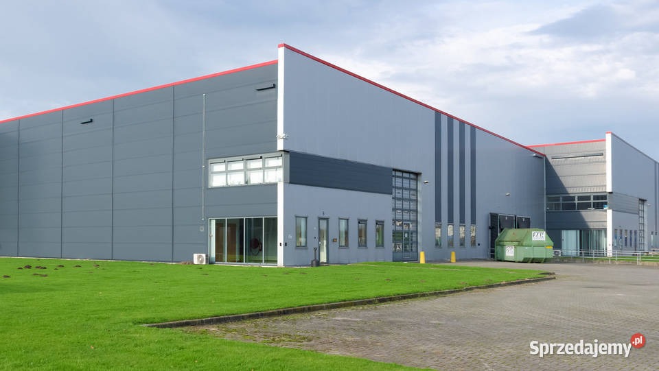 Nowoczesny budynek magazynowy z szarą elewacją panelową i czerwonymi akcentami, widok z zewnątrz, z zielonym trawnikiem i kontenerem na pierwszym planie, pochmurne niebo.